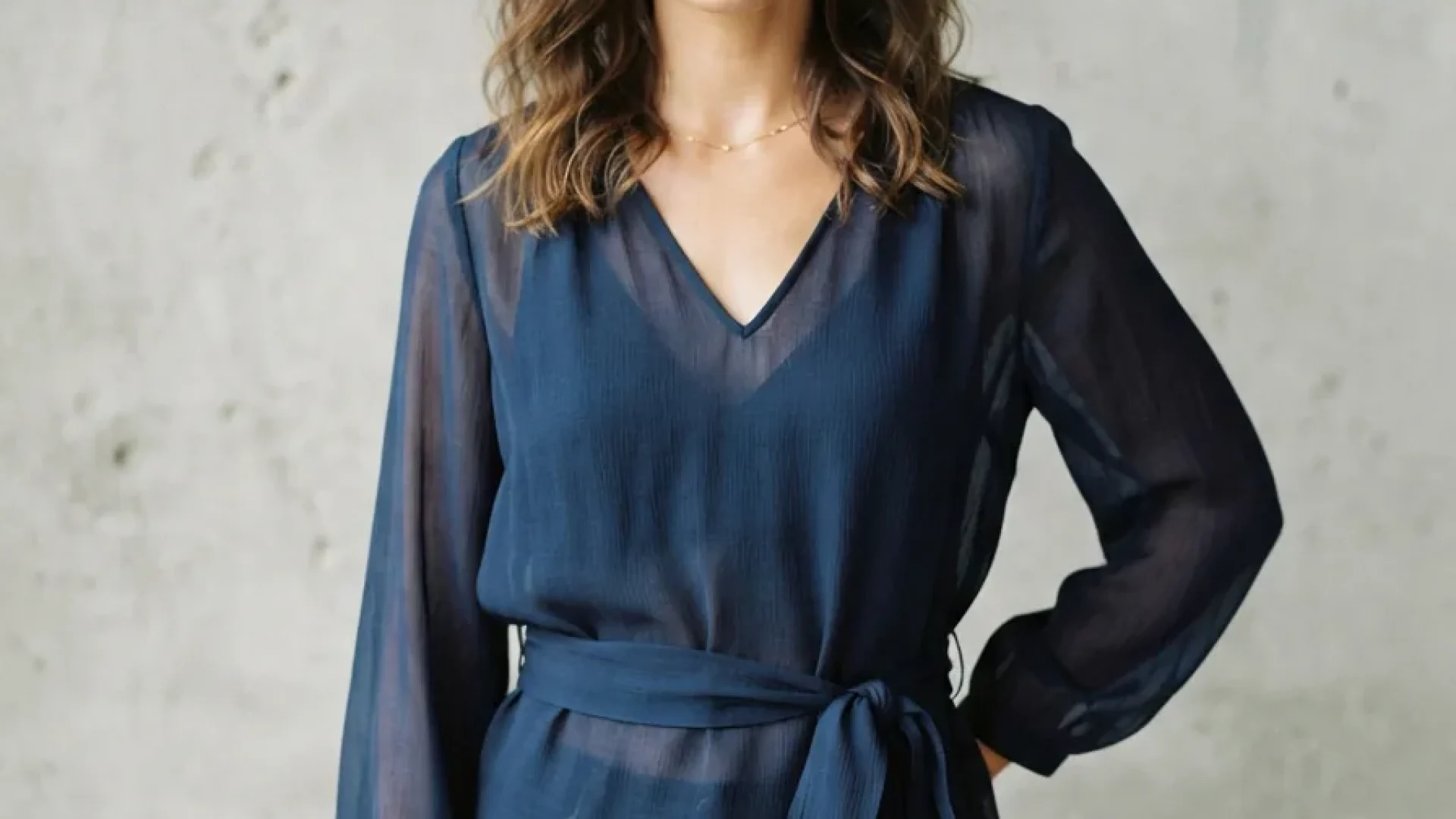 A model confidently wearing a sheer navy Arden B blouse with a tie waist, standing against a neutral, textured wall.